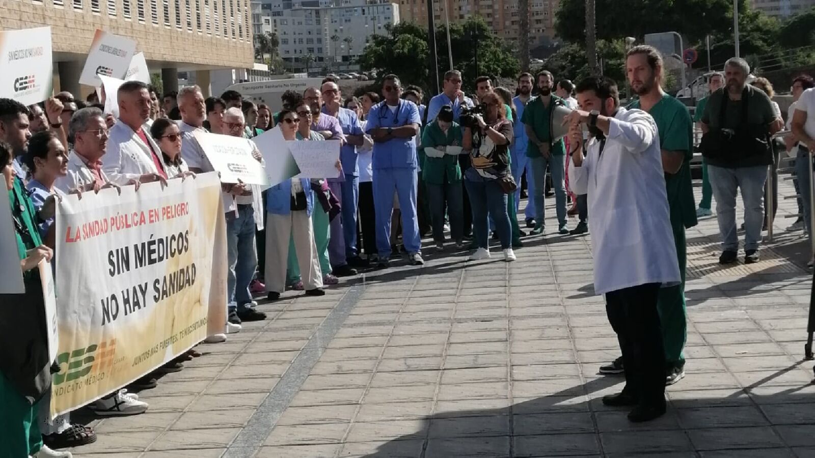 Un médico con bata blanca habla con un megáfono ante compañeros que sostienen pancartas