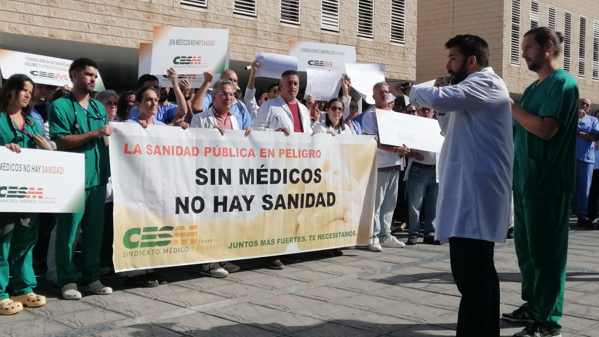 Concentración de médicos al sol frente a un hospital: sostienen una gran pancarta con la frase «Sin médicos no hay sanidad» y carteleras de «¡Condiciones laborales dignas!»