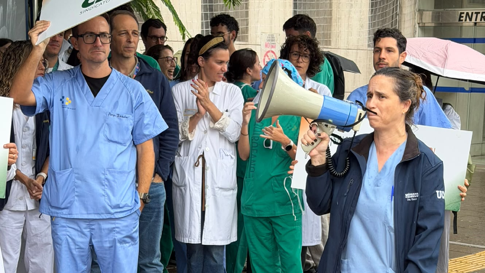Una médica con pijama azul habla con un megáfono bajo la lluvia, rodeada de compañeros con paraguas y pancartas