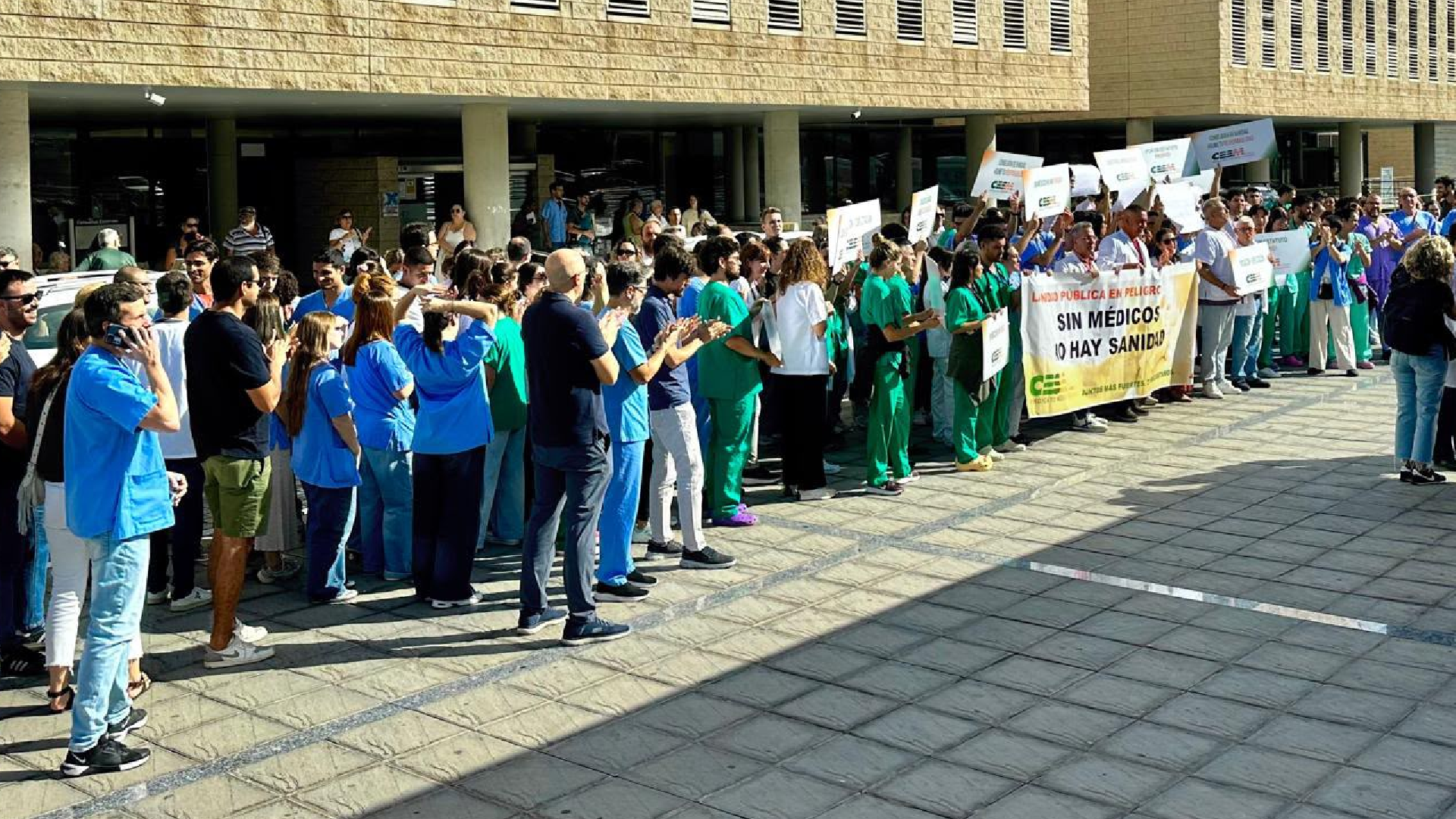 Gran círculo de médicos con batas y pijamas azules y verdes aplaudiendo frente a la fachada del hospital, con pancartas en las manos